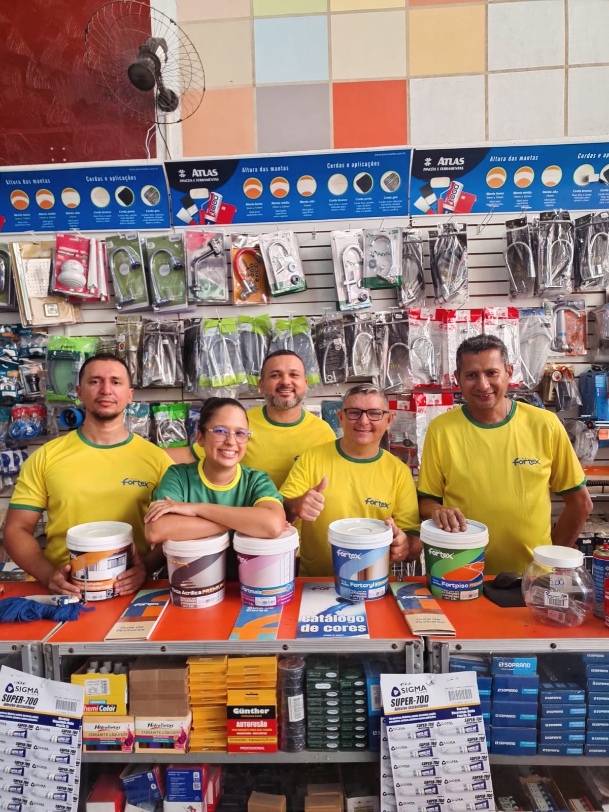 Equipe da Casa Soares atrás do balcão, com latas de tinta, guias de cores e o painel de metais e ferragens ao fundo.